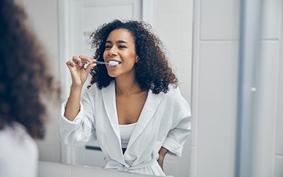 A woman with curly hair brushing her teeth in front of a bathroom mirror